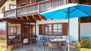 Een houten huis met twee verdiepingen, een balkon, een terras met tafel en stoelen, en een blauwe parasol.