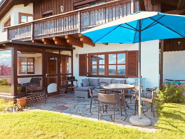 Une maison en bois à deux étages, dotée d’un balcon, d’une terrasse avec une table et des chaises, et d’un parasol bleu.