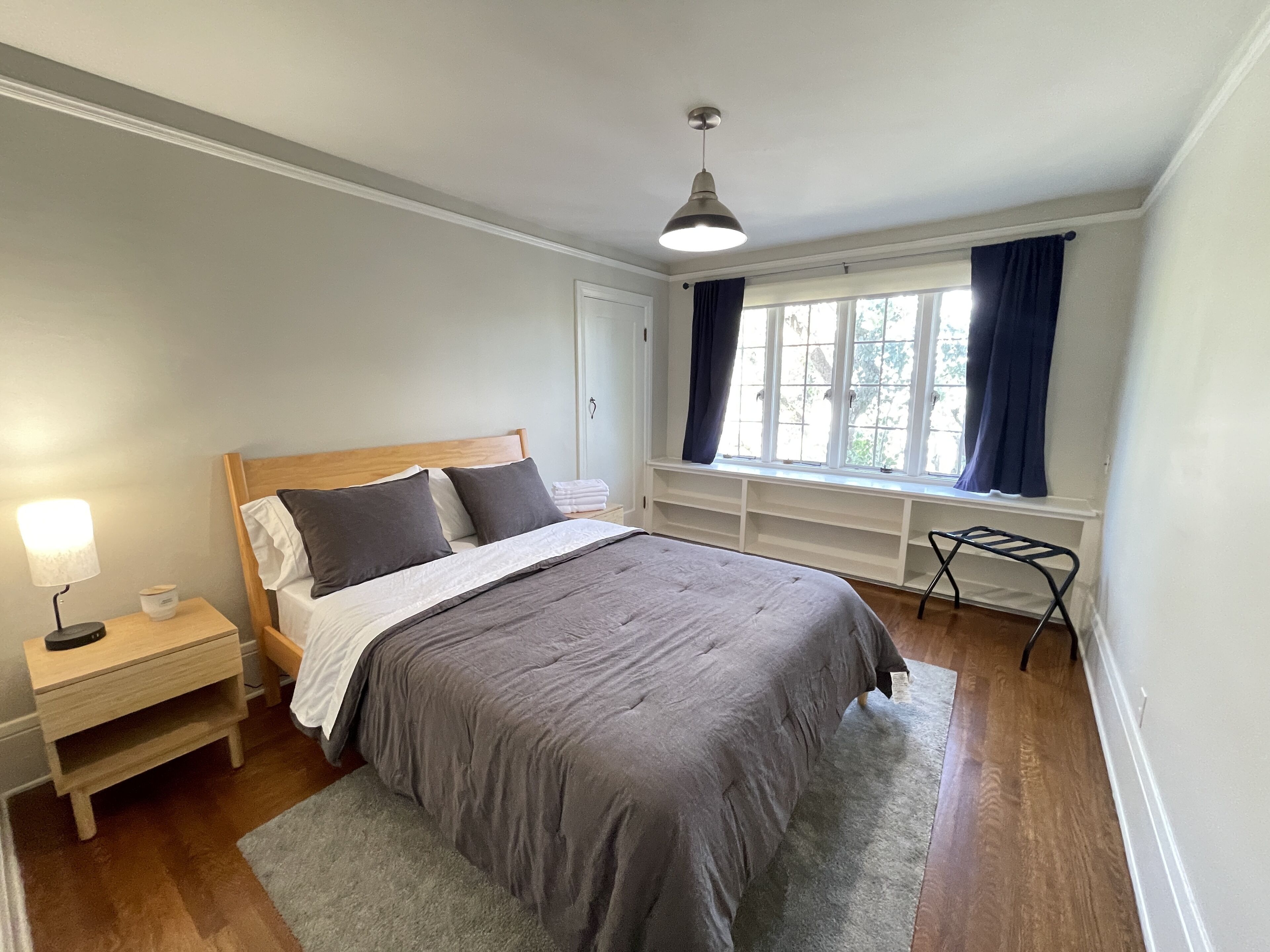 A bedroom with a wooden bed, a bedside table with a lamp, a window with curtains, and a built-in shelf.