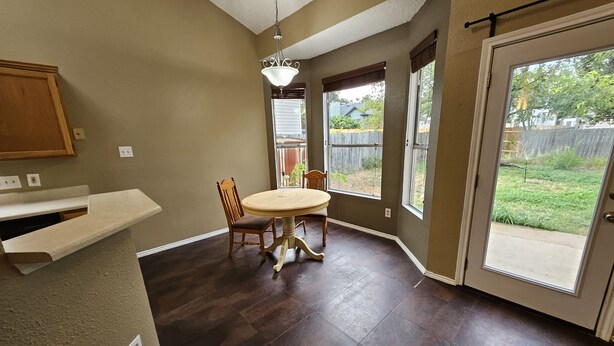 A kitchen with a wooden table and chairs, a hanging light fixture, and a view of the backyard through large windows.