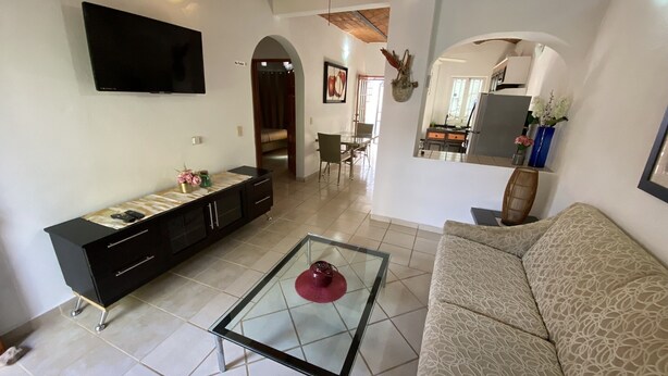 Una sala de estar con un sofá, una mesa de centro de vidrio, un televisor montado en la pared, un mueble con objetos decorativos y una zona de comedor al fondo.
