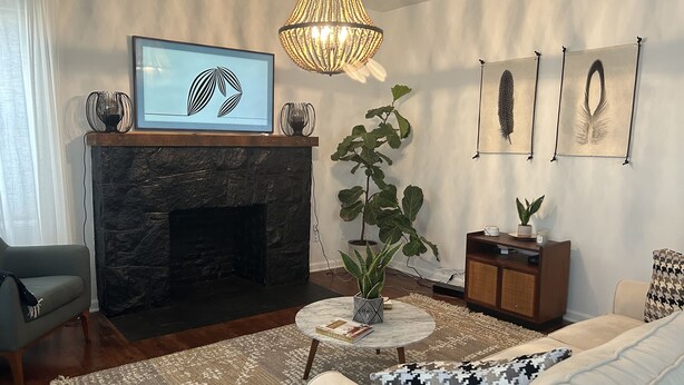 Una sala de estar con chimenea de piedra, una planta grande en maceta, una mesa de centro redonda y un sofá con cojines estampados.