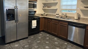 Una cocina con un refrigerador de acero inoxidable, un microondas, una estufa y un lavavajillas. Hay gabinetes de madera, una encimera de granito y un piso con diseño.