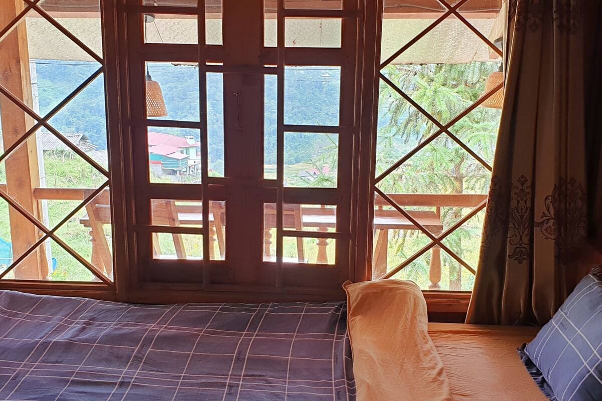A room with a bed, a window with a wooden lattice design, and a view of greenery and buildings outside.