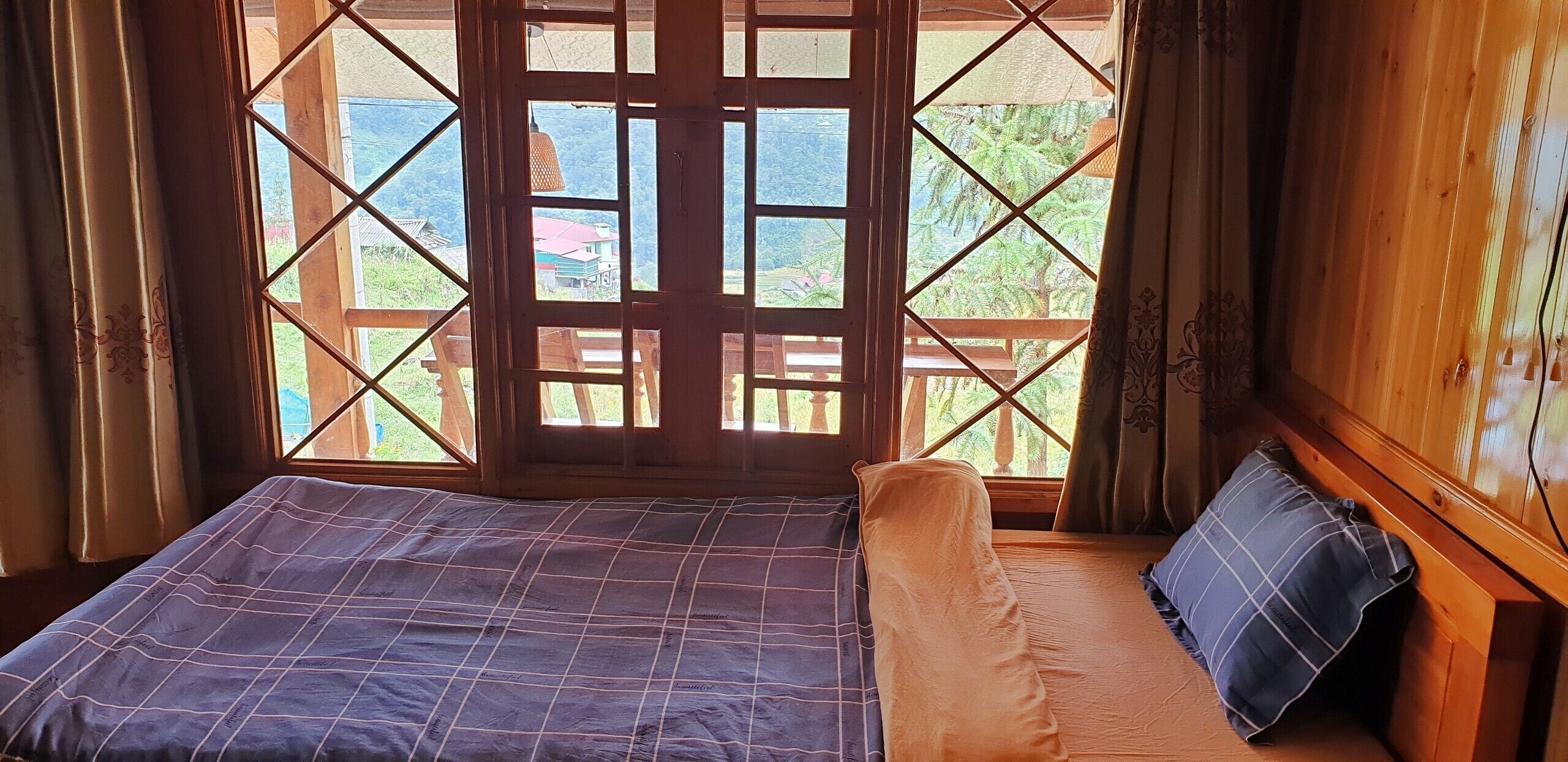 Una habitación con cama, ventana que ofrece vistas a follaje y un edificio, y paredes de madera.