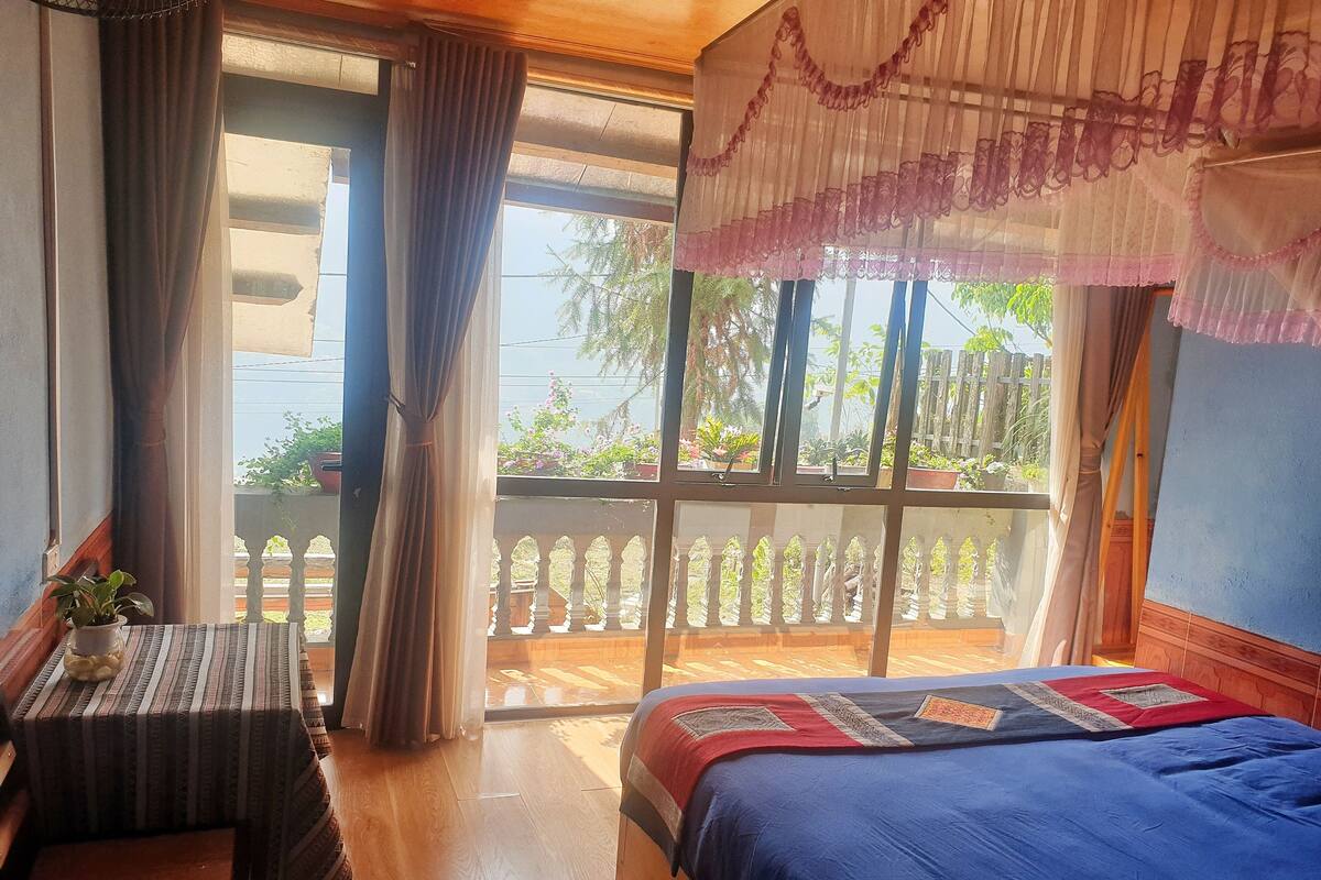 A room with a bed, a table, and a view of a balcony with plants.