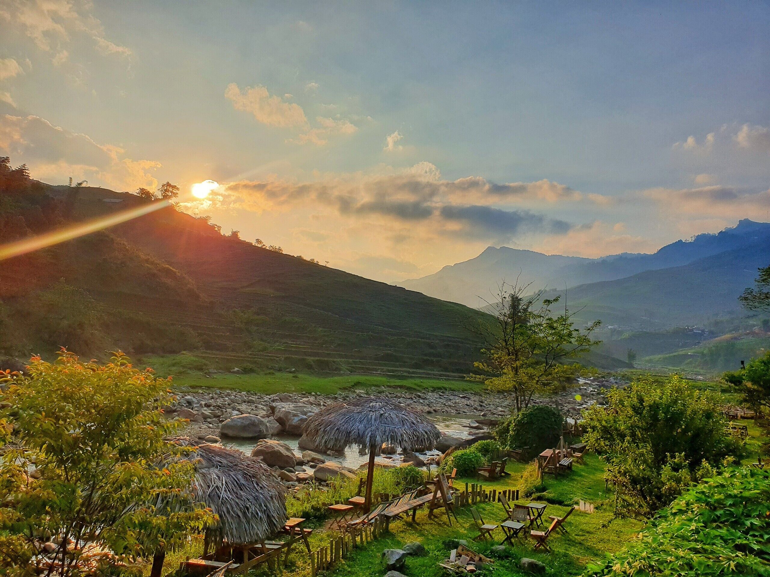 A scenic outdoor area with a river, wooden seating, and thatched structures.
