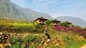A rural setting with traditional thatched-roof houses, a stone wall, and a person sitting on the wall. - Peace Home (Ta Van)