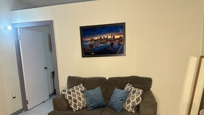 Een woonkamer met een bruine bank, twee gekleurde kussens en een stadsgezicht in een lijst aan de muur.