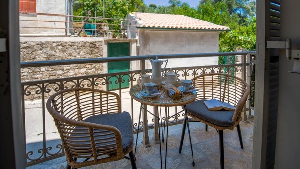 A balcony with wicker chairs and a small table, overlooking a residential area with stone walls and a tiled roof.