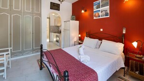 A bedroom with a bed, a wardrobe, a kitchen visible through an archway, and a red wall with a framed picture.