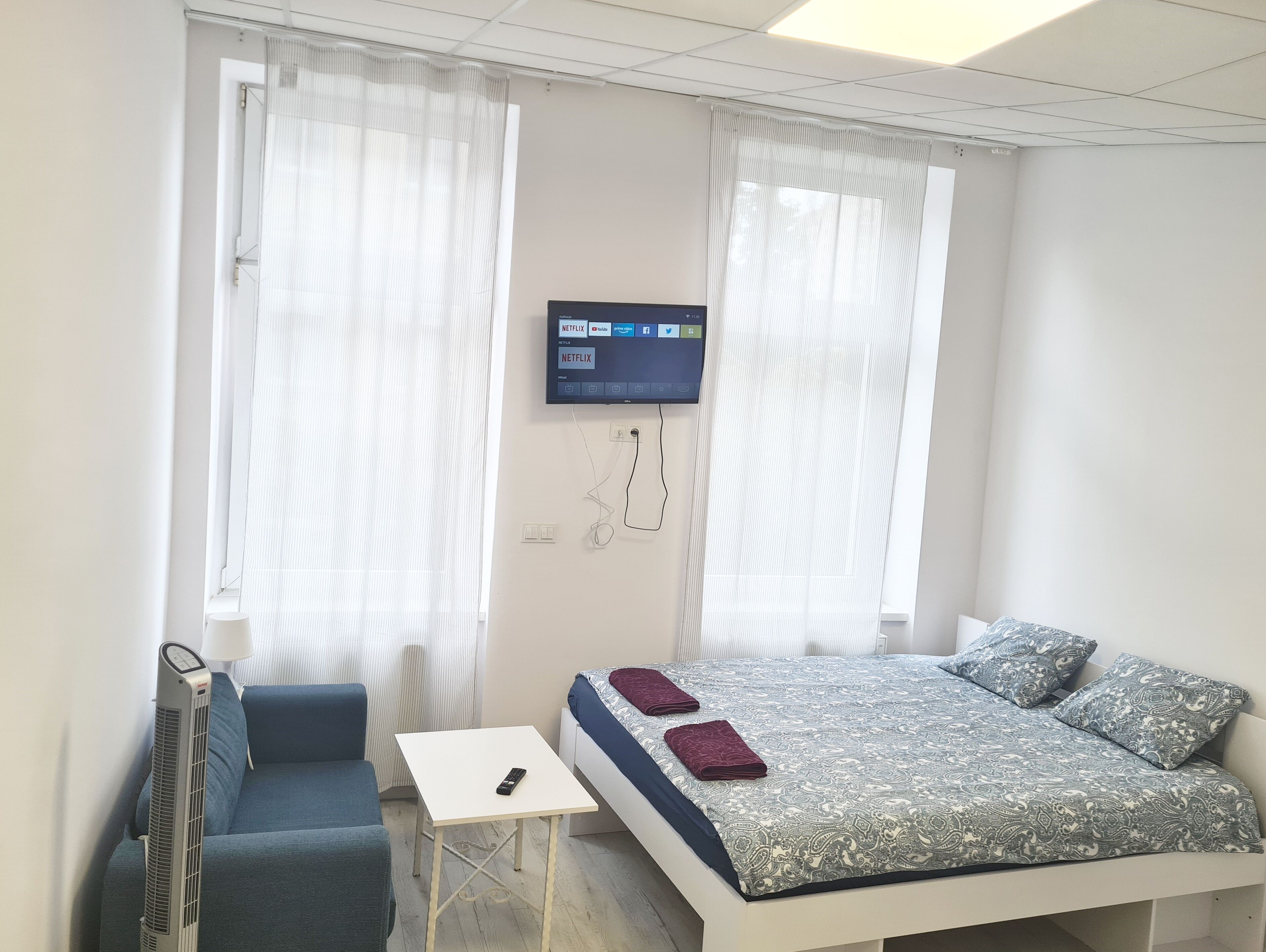 A bedroom with a bed, a small table, a chair, a TV mounted on the wall, and a standing heater.