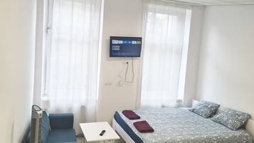 A bedroom with a bed, a small table, a chair, a TV mounted on the wall, and a standing heater.
