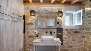 Un baño con pared de piedra, vigas de madera, un espejo grande y un lavamanos blanco.