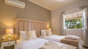 Ein Hotelzimmer mit zwei Betten, einem Fenster mit Blick auf Bäume, einem Nachttisch mit Lampe und einer Wand-Klimaanlage.