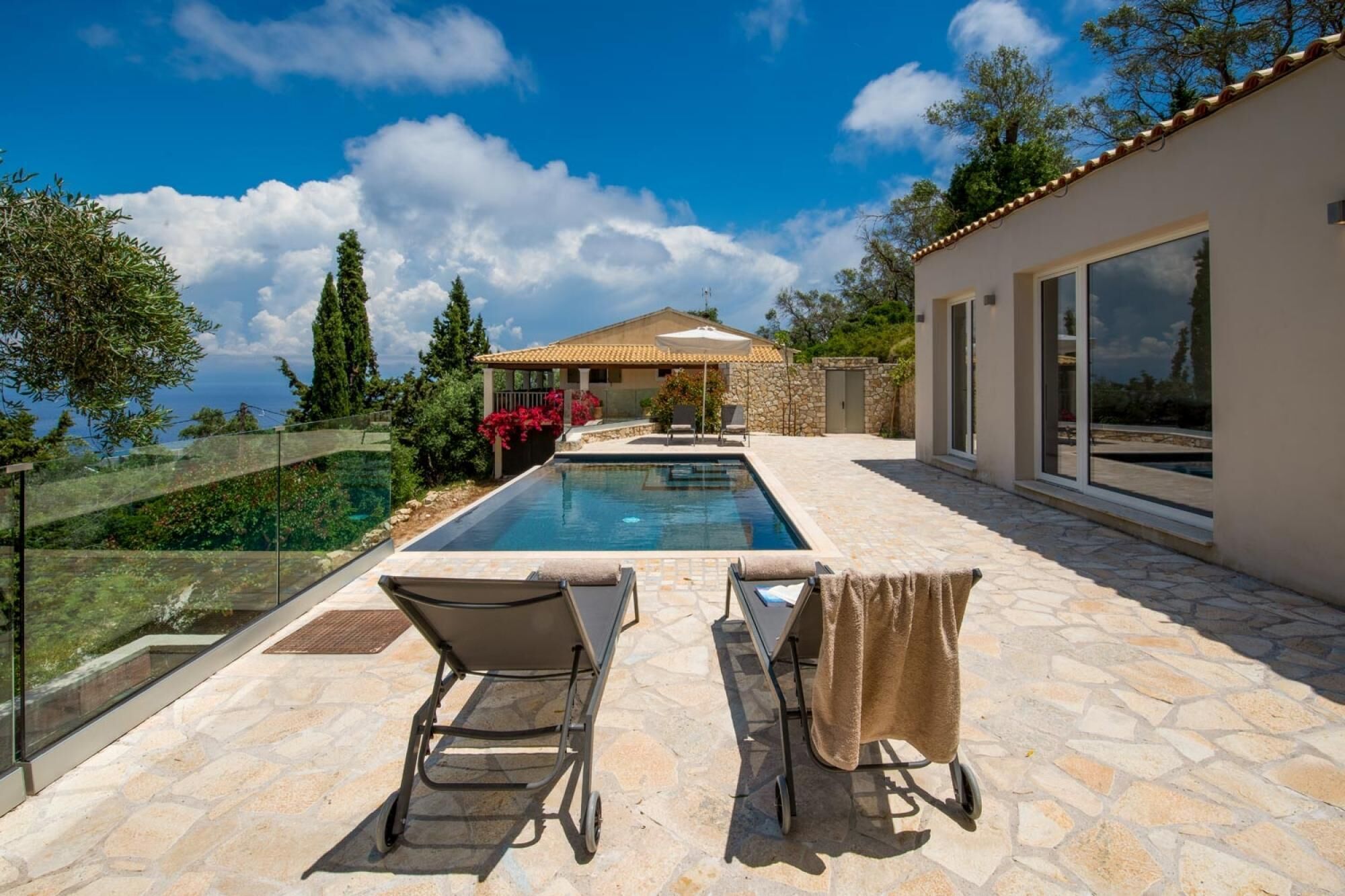 Ein Poolbereich mit zwei Liegestühlen und einem Tisch, eine Steinterrasse, ein Haus mit Ziegeldach und ein klarer, blauer Himmel.
