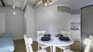 Un coin repas avec une table blanche dressée pour quatre personnes, entourée de chaises blanches, dans une pièce moderne aux poutres apparentes et avec une cuisine en arrière-plan.