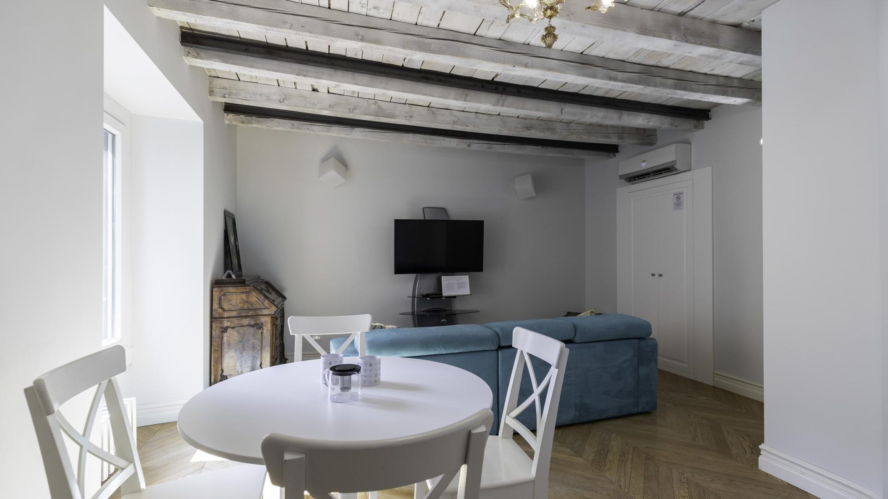 A room with a white round table, two white chairs, a blue sofa, a flat-screen TV mounted on the wall, and a wooden cabinet.