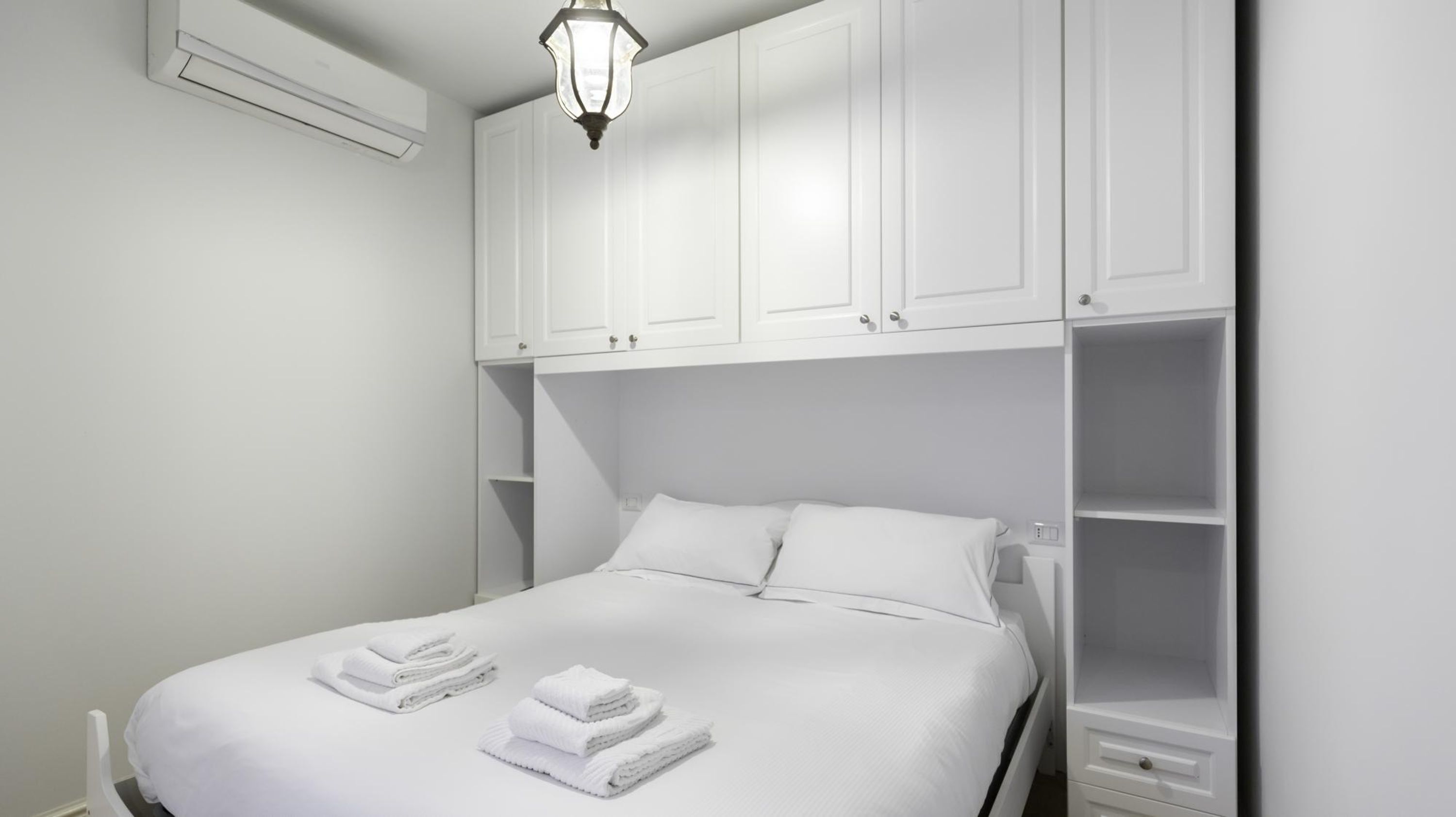 Een net opgemaakt bed met witte lakens en handdoeken, een witte kledingkast met meerdere deuren, en een aan de muur bevestigde airconditioning.'
