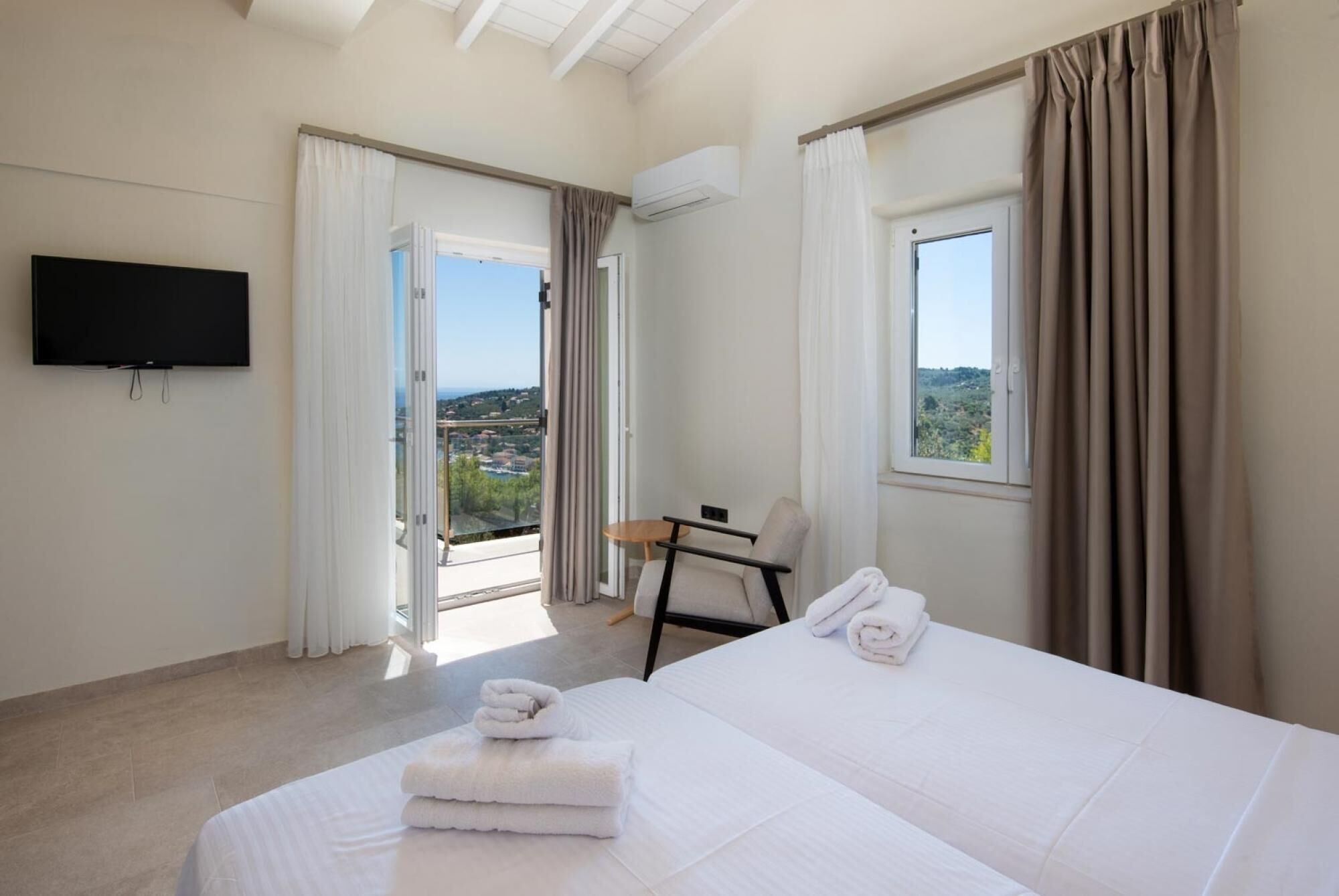 Ein Hotelzimmer mit Bett, Sessel, Fernseher und einem Balkon mit Vorhängen.