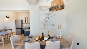 A modern dining area with a wooden table set for four, surrounded by tufted chairs. A large abstract painting hangs above the table, and a rustic chandelier is visible.