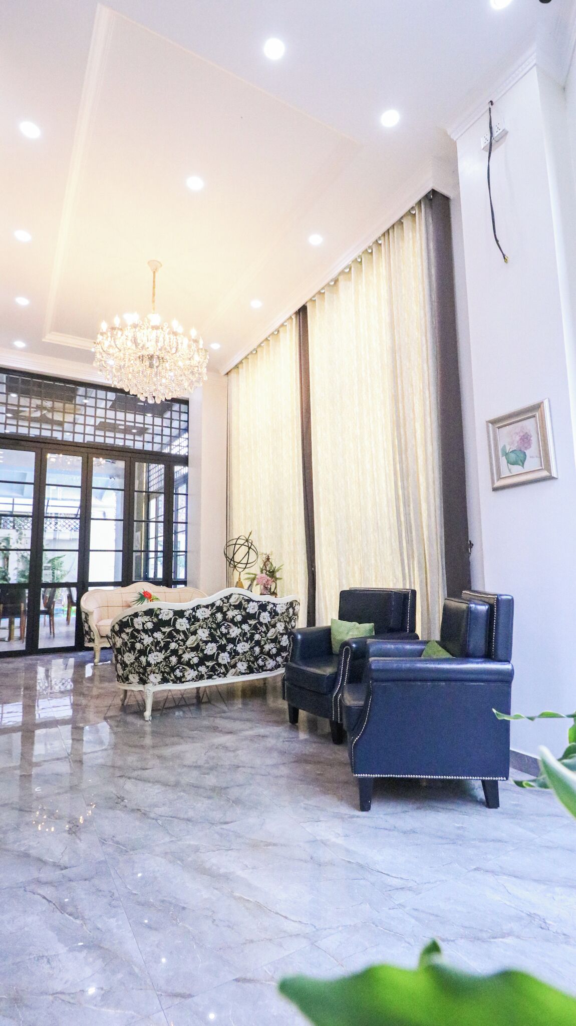 A spacious living room with a chandelier, a sofa, and a blue armchair. There is a large window with curtains, and a plant is visible in the corner.