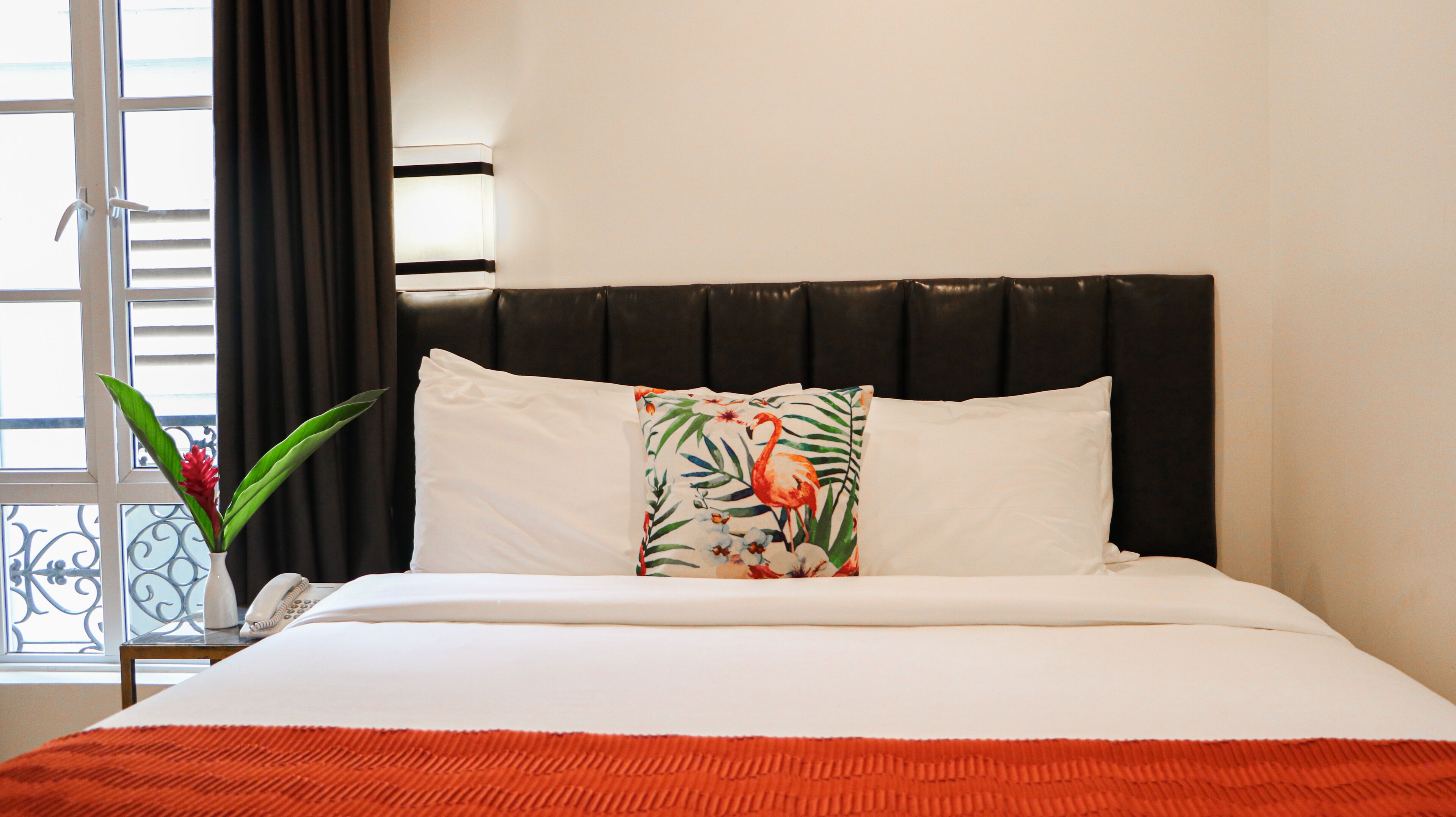 Une chambre d’hôtel avec un lit, une fenêtre avec des rideaux, une plante et un coussin décoratif.