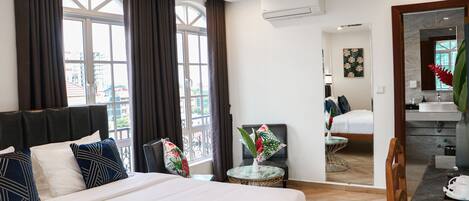 Habitación de hotel con una cama grande, una mesa redonda con un florero, una silla, vista de la ciudad a través de grandes ventanales y un baño con lavabo y espejo.