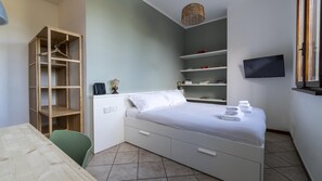Ett sovrum med en säng, ett skrivbord, en stol, en tv och hyllor med böcker och prydnadsföremål.