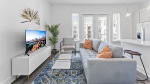 A modern living room with a sofa, a coffee table, and a TV. There are windows with blinds, a plant, and a decorative wall piece. - Chic West Downtown Studio w Washer Dryer (Austin)