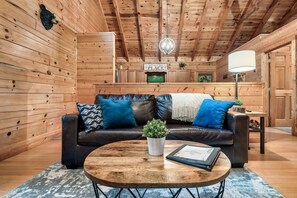 Interior de uma cabana de madeira com sofá de couro, mesa de centro redonda, planta em vaso e uma placa com a inscrição 'Bem-vindos ao Nosso Lugar'.