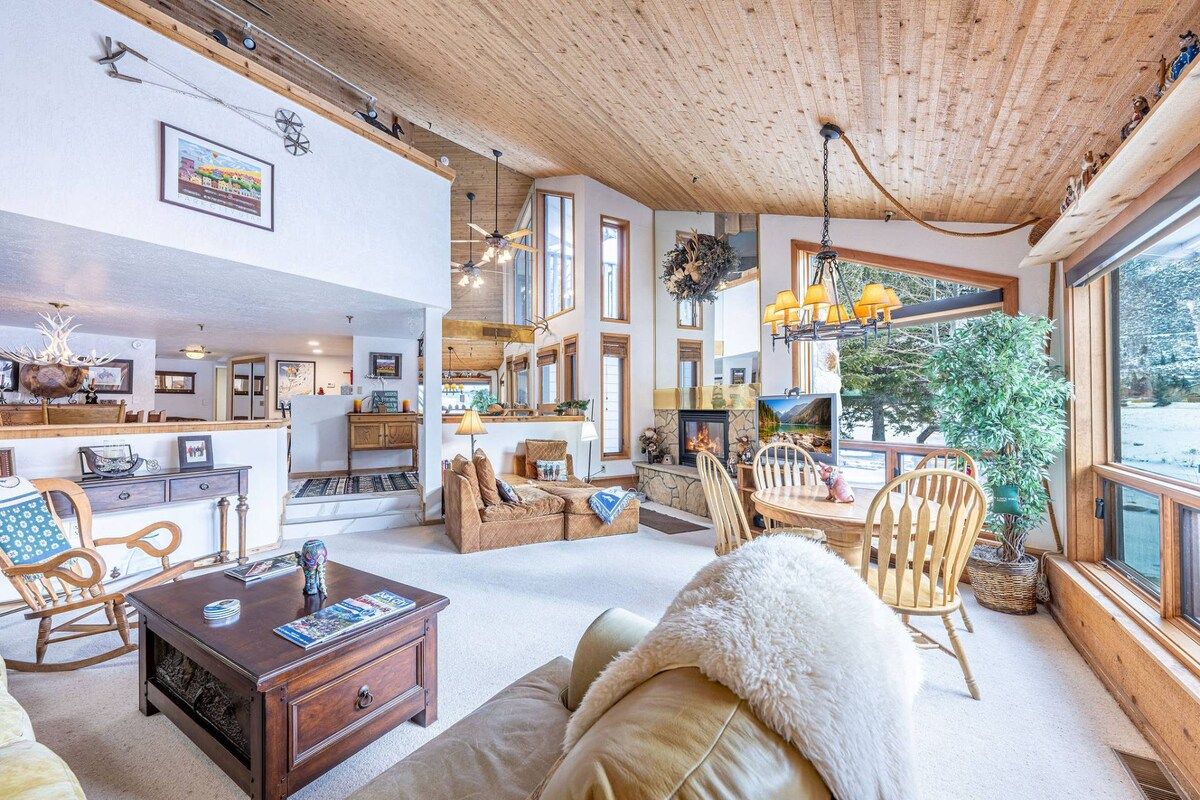 Amplia sala de estar con chimenea, techo de madera y ventanas grandes que ofrecen vistas al exterior.