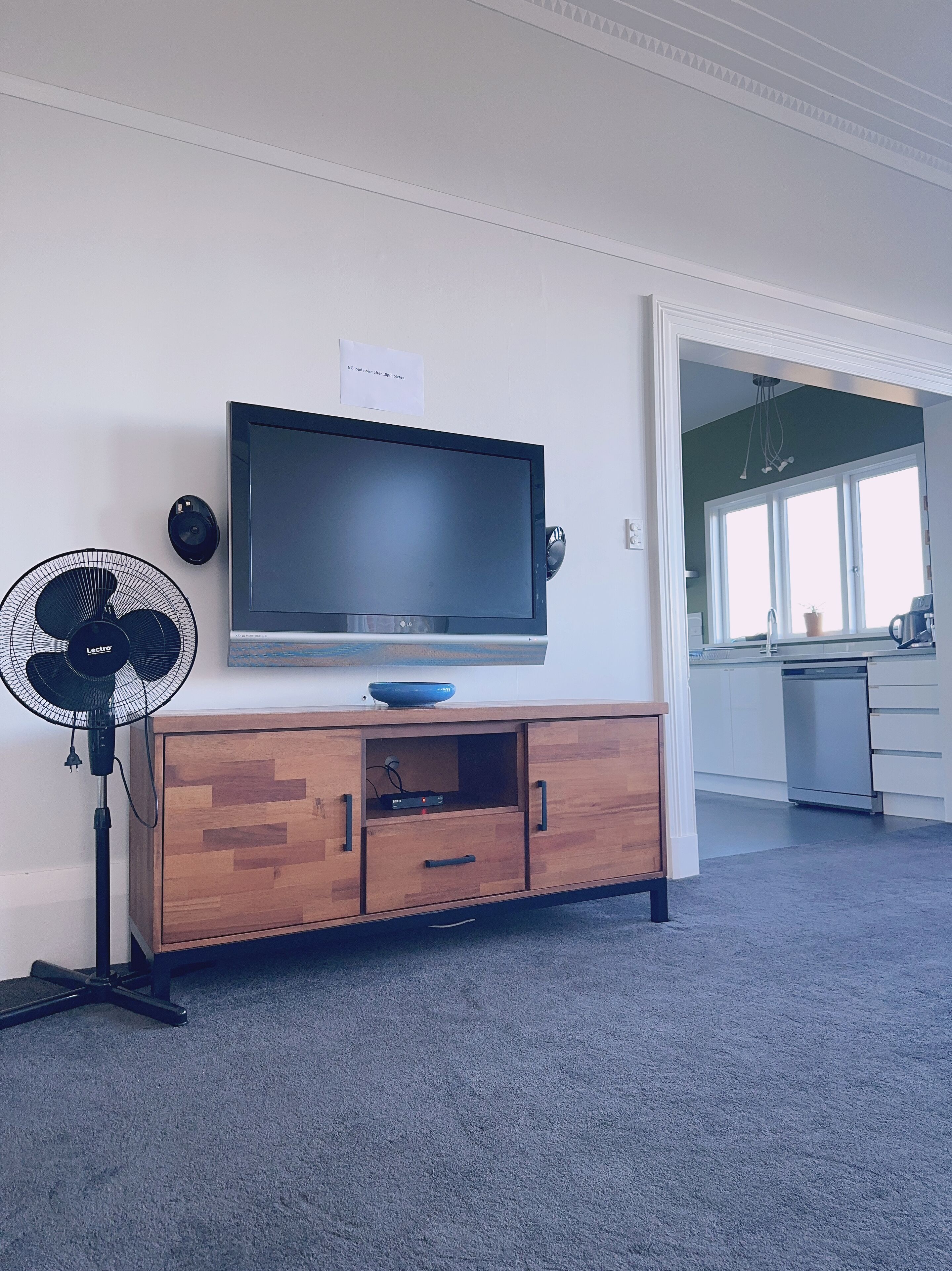 Uma sala de estar com uma TV de tela plana em um armário de madeira, um ventilador e uma cozinha visível através de uma porta aberta.