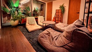 A cozy living room with a brick wall, large windows, and a variety of plants. There are two sofas, a small round table, and a guitar in the background. - HHZib - homestay on a peaceful hill (Seoul)