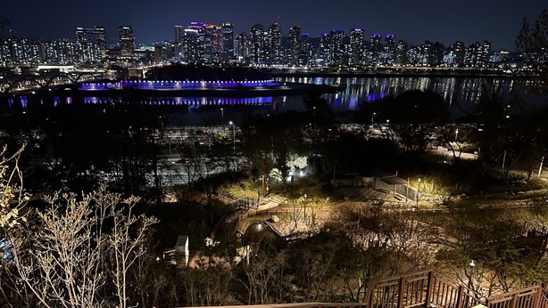 A nighttime view of a city skyline reflected in a body of water, with a wooden deck and railing in the foreground. - HHZib - homestay on a peaceful hill (Seoul)