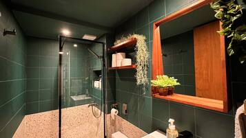 A modern bathroom with dark green tiles, a glass shower enclosure, a white sink, and a wooden shelf with plants.