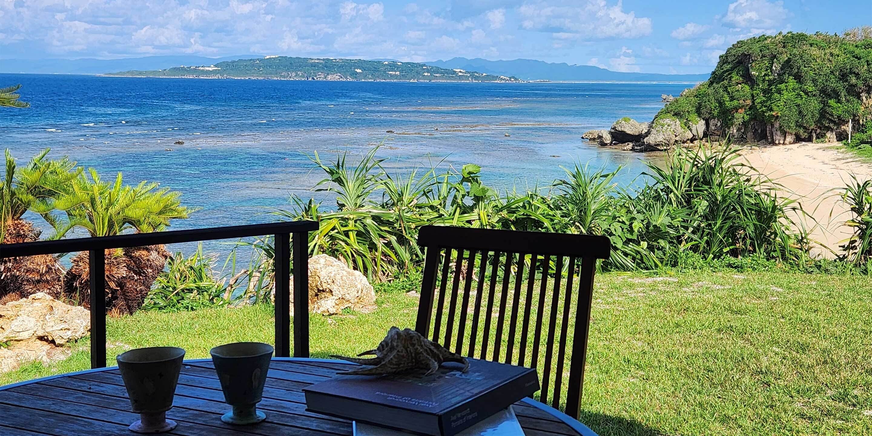 A wooden deck with a table, two cups, and a book. The deck overlooks a beach with clear blue water and distant land.