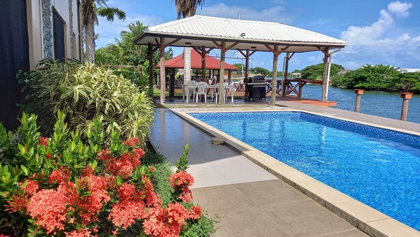 Uma piscina com deck ladrilhado, rodeada por vegetação exuberante e um gazebo com mobiliário para área externa.
