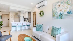 A modern living room with a white sofa, teal cushions, a wooden coffee table, and a kitchen in the background.