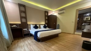 A modern hotel room with a wooden floor, a bed with white bedding and dark pillows, a wooden wardrobe, a small bedside table with a lamp, and a bathroom visible through an open door.