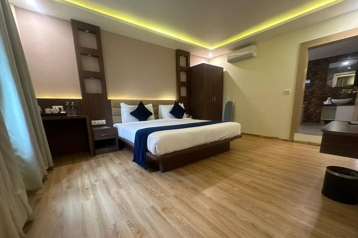 Una habitación de hotel moderna con piso de madera, una cama con ropa blanca y almohadas oscuras, un armario de madera, una mesita de noche pequeña con una lámpara, y un baño visible a través de una puerta abierta.