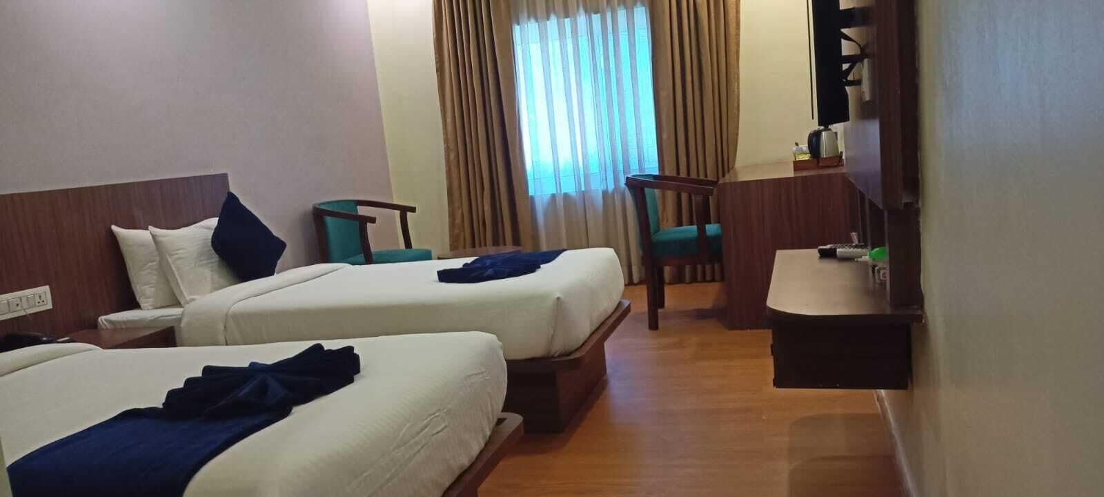 A hotel room with two beds, a small desk, a chair, and a window with curtains.