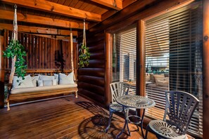 A wooden deck with a swing, a table, and chairs, and a view through large windows with blinds. - Smoky Mtn Fall Foliage Cabin – Hot Tub, Indoor Pool, Near Dollywood, Sleeps 6 (Sevierville)