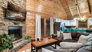 L’intérieur d’un chalet accueillant, avec une cheminée en pierre, des murs en bois, un coin salon doté d’un canapé et de coussins, et une cuisine à l’arrière-plan.