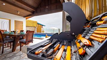 A barbecue grill with tools, a dining table with chairs, and a view of a pool and beach.