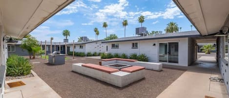 A modern outdoor patio with a fire pit, seating, and a view of palm trees and a building in the background.