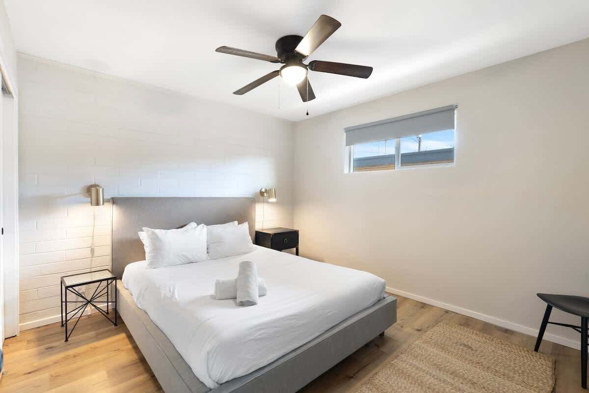 Une chambre à coucher avec un lit, un ventilateur de plafond, une table de chevet, une fenêtre et une petite table.