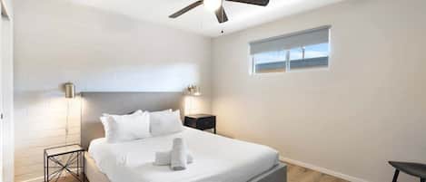 Une chambre à coucher avec un lit, un ventilateur de plafond, une table de chevet, une fenêtre et une petite table.
