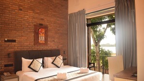 Hotelkamer met een bakstenen muur, een groot bed met witte beddengoed, een houten hoofdbord, een nachtkastje met een telefoon en uitzicht op een balkon met een boom.
