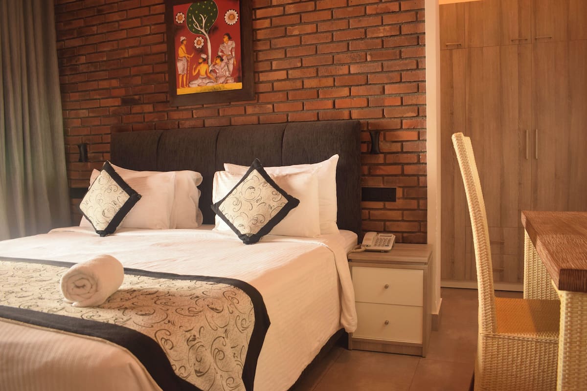 A hotel room with a brick wall, a bed with a patterned bedspread, two pillows with a black border, a bedside table with a phone, and a wicker chair.